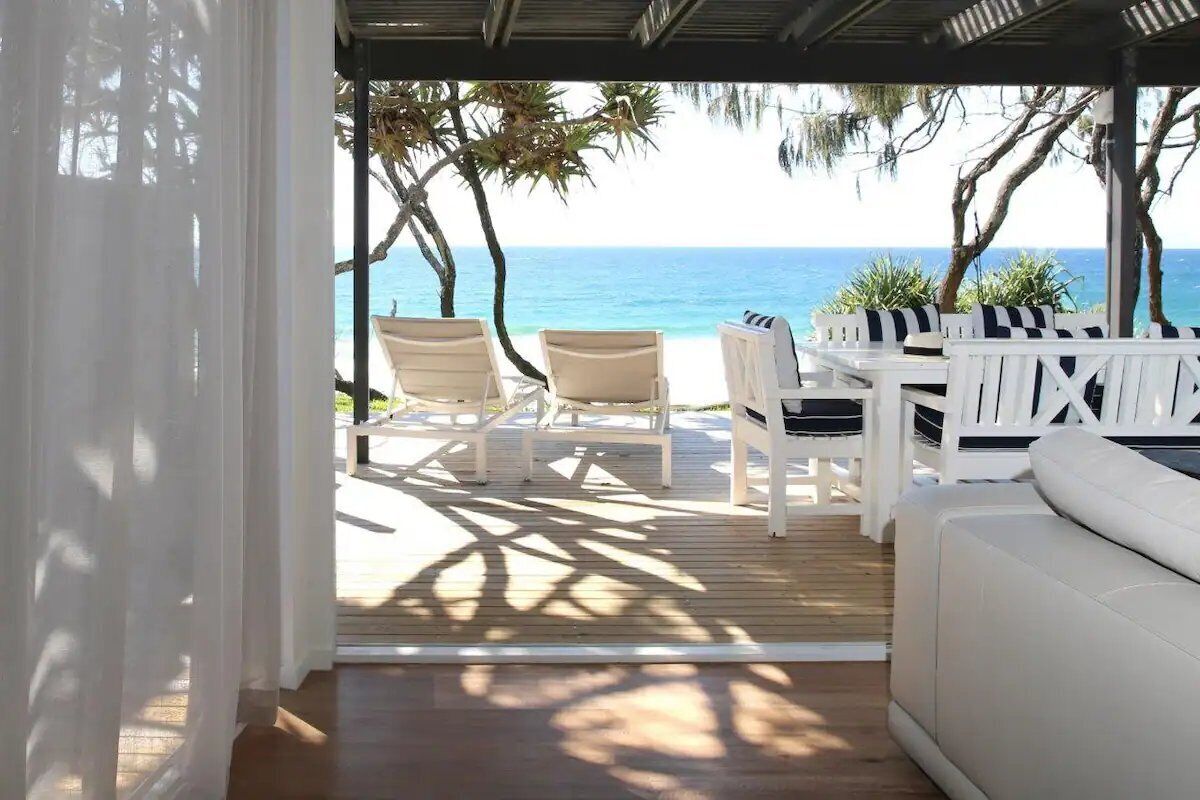 A covered outdoor seating area with a view of the ocean, wooden deck, and white furniture.