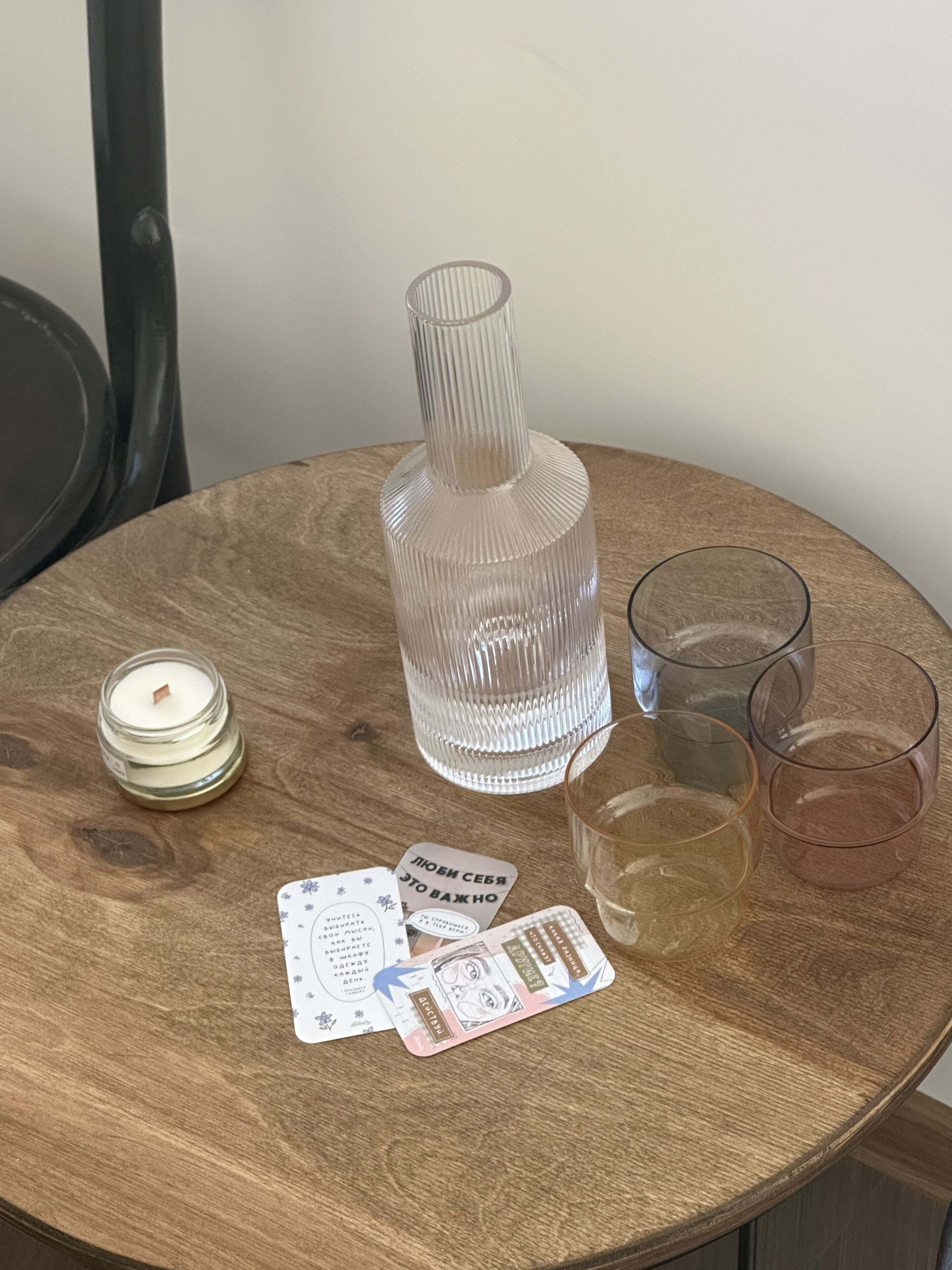 A round wooden table with a glass candle, a clear glass bottle, two glasses, and a few cards.