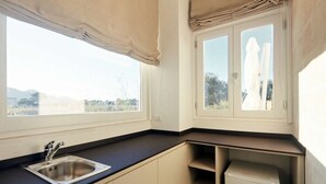 A kitchen with a sink, cabinets, and a refrigerator. There are windows with curtains and a view of the outdoors. - Cala Monaci Apartment (Favignana)