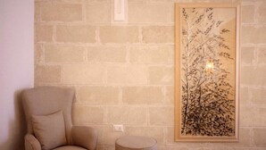 A room with a beige brick wall, a framed botanical print, a beige armchair, and a round ottoman. - Cala Monaci Apartment (Favignana)