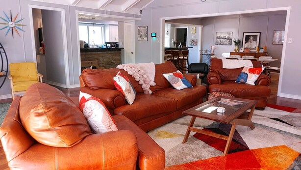 A living room with brown leather sofas, a coffee table, and a colorful rug. There is a dining area in the background with a dining table and chairs. - Mid Century Modern Nights Dream (Houston)
