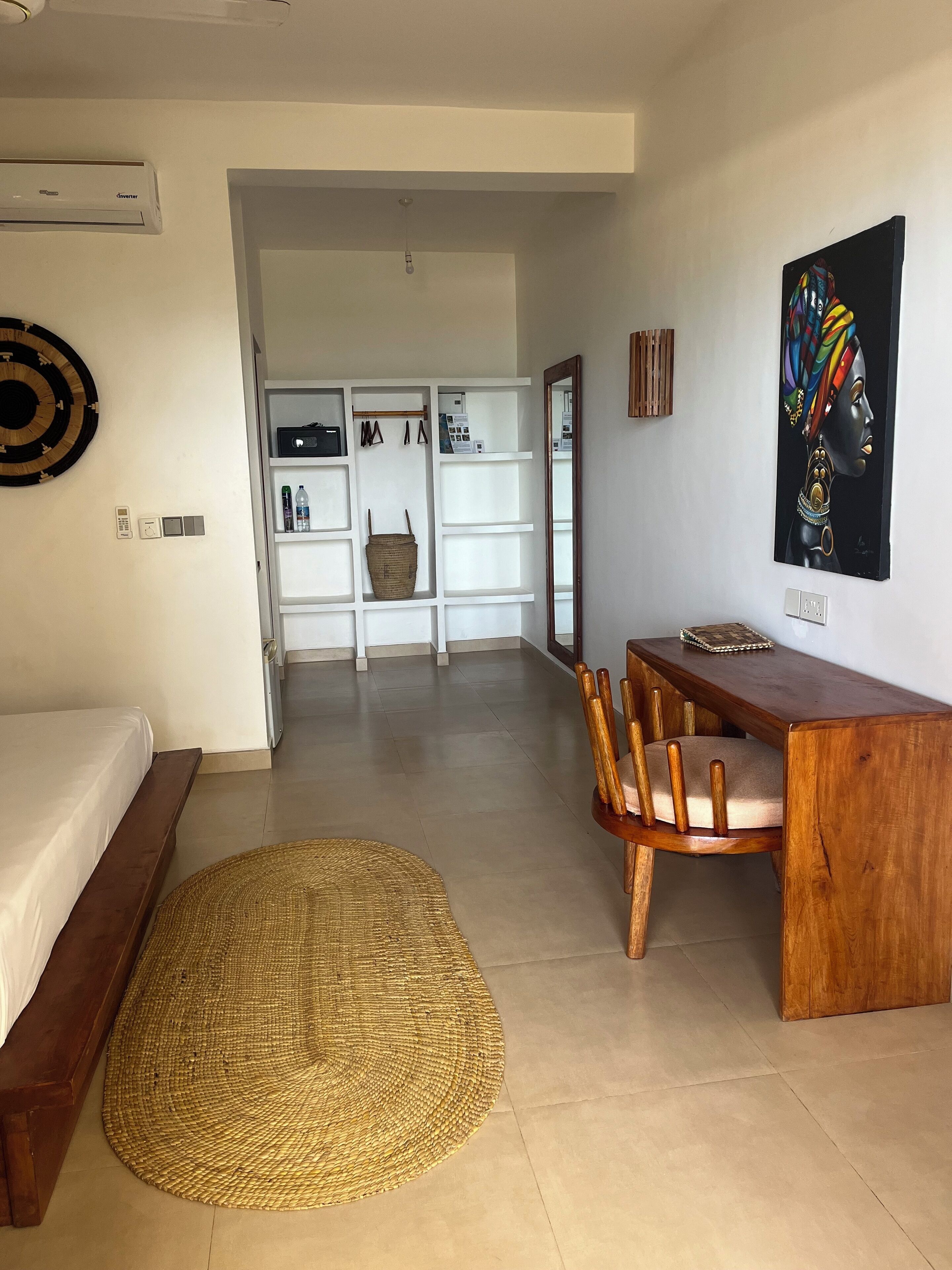 A modern hotel room with a bed, a wooden desk, a woven rug, a white wardrobe, and a painting on the wall.