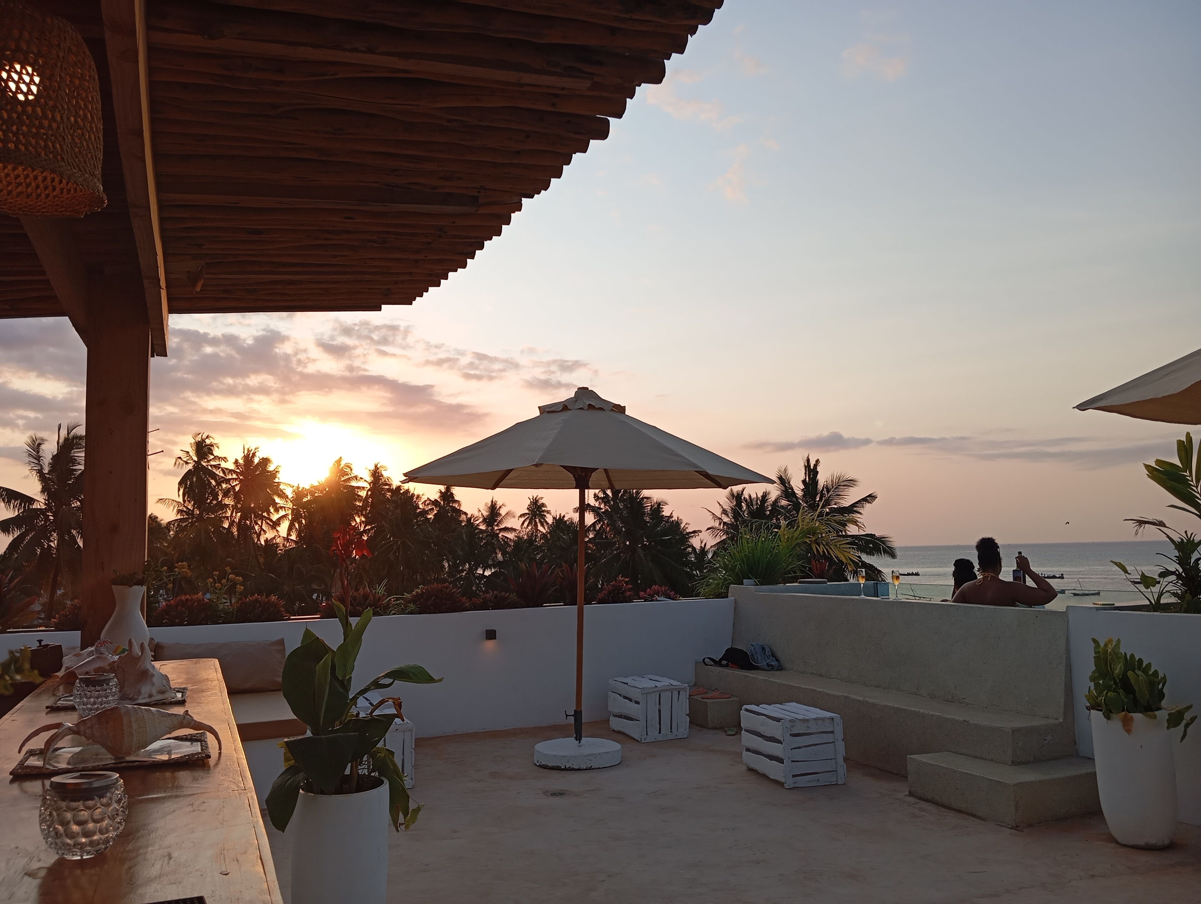 Een terras met uitzicht op een zonsondergang, palmbomen en een strand.