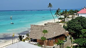 A beach view with a thatched-roof structure, palm trees, and a clear blue sea. - Aluna Beach Apartments (Nungwi)
