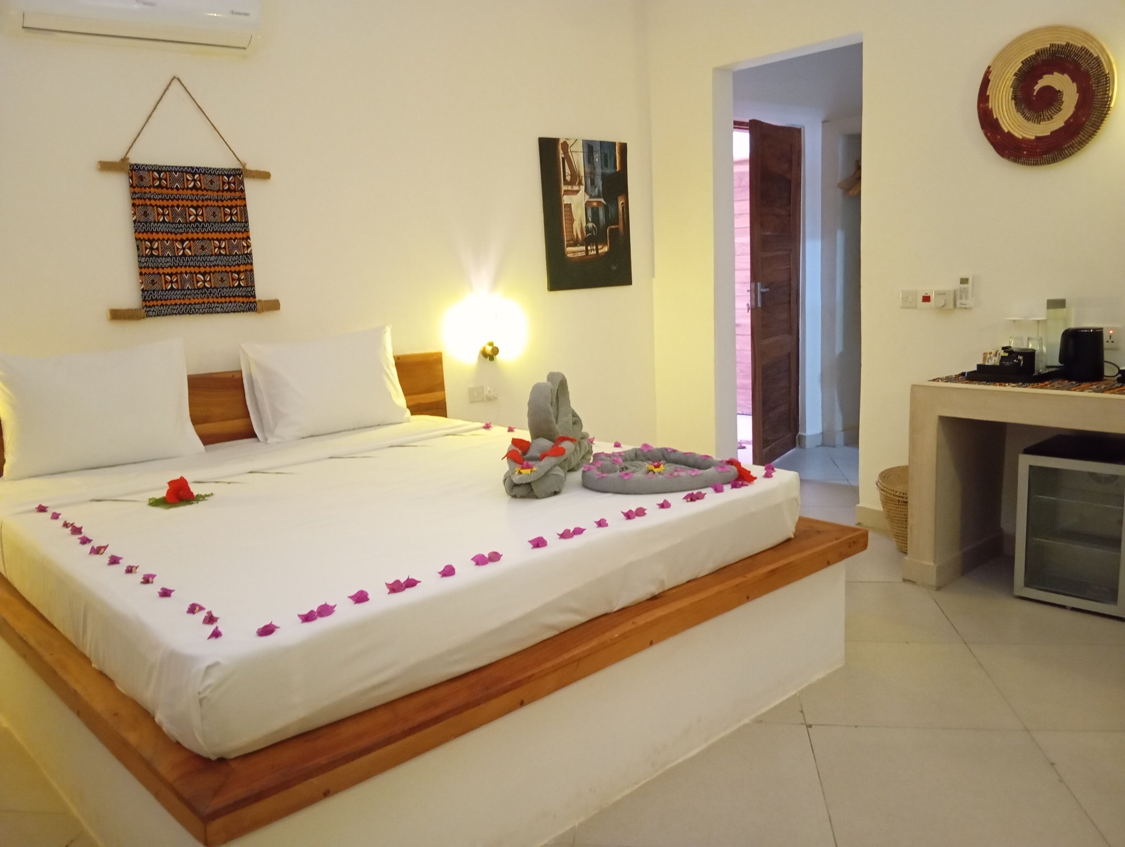 A neatly made bed with white linens, a gray towel, and pink flower petals. There is a wall-mounted artwork above the bed and a small table with a coffee machine to the right.