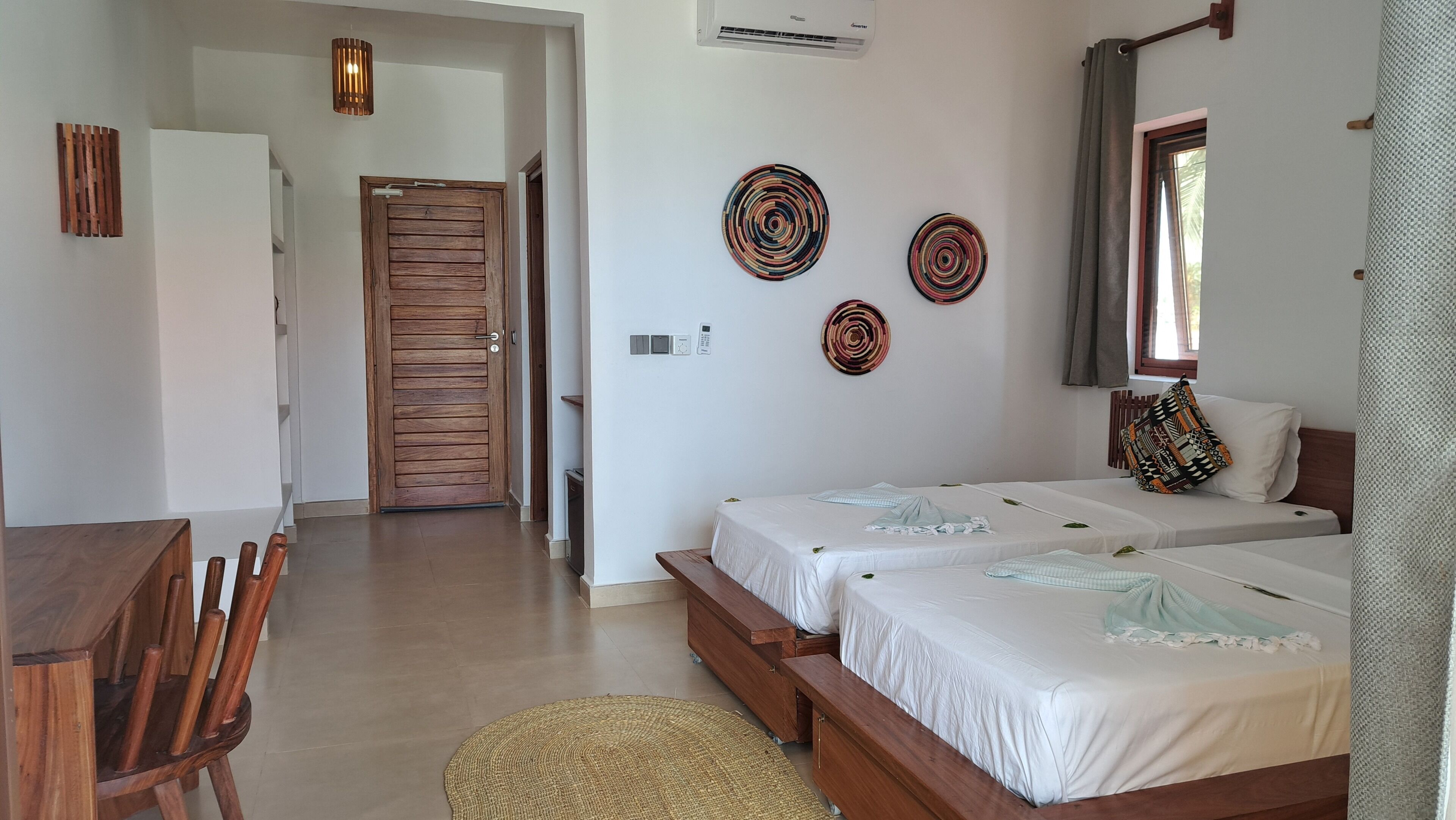 A hotel room with two beds, a wooden door, a small desk, and a woven mat.