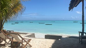 A beach with clear turquoise water, several boats, and wooden lounge chairs under a palm tree. - Aluna Beach Apartments (Nungwi)