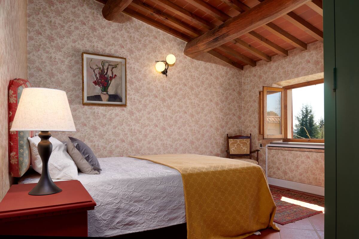 Une chambre à coucher avec un lit, une table de chevet avec une lampe, un tableau au mur et une fenêtre donnant sur des arbres.