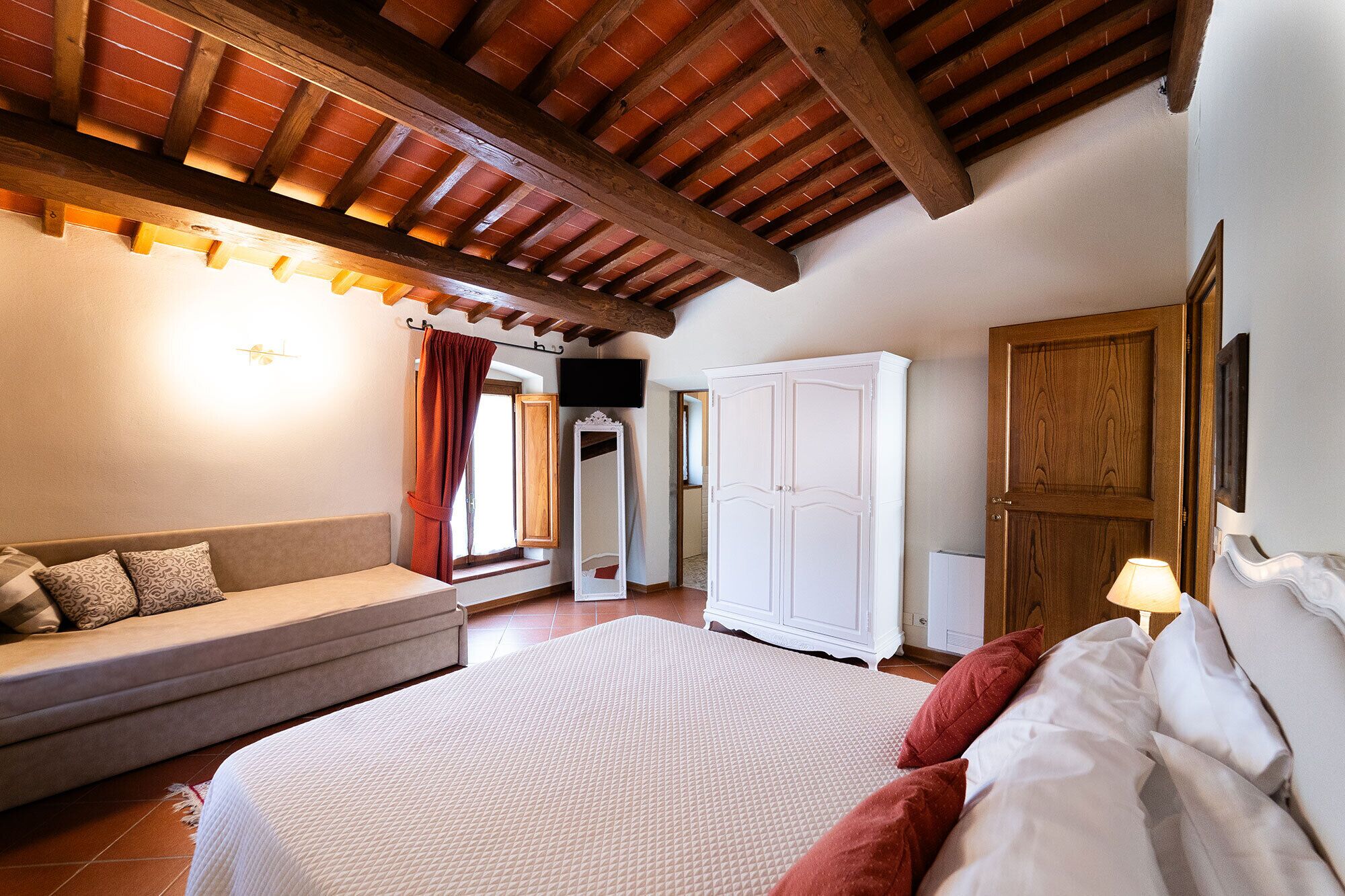 A bedroom with a bed, a sofa, a wardrobe, and a ceiling with exposed wooden beams.
