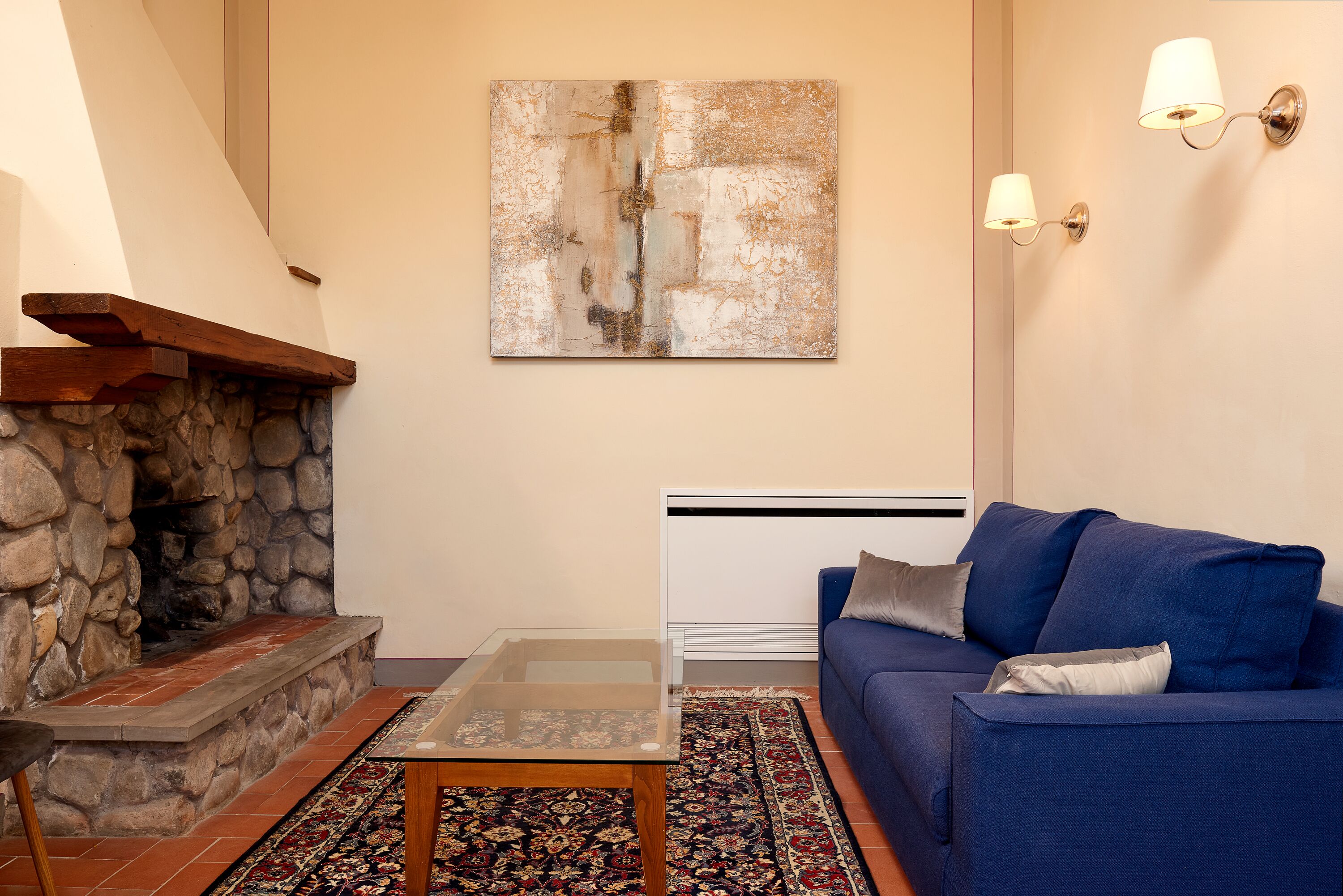 Un salon avec une cheminée en pierre, un canapé bleu, une table basse en verre et un grand tableau abstrait accroché au mur.
