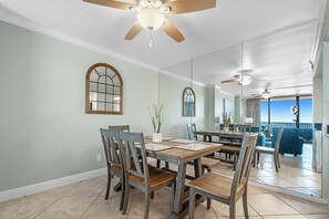 Un espace repas avec une table et des chaises en bois, un ventilateur de plafond et une vue sur l’extérieur grâce à de grandes fenêtres.