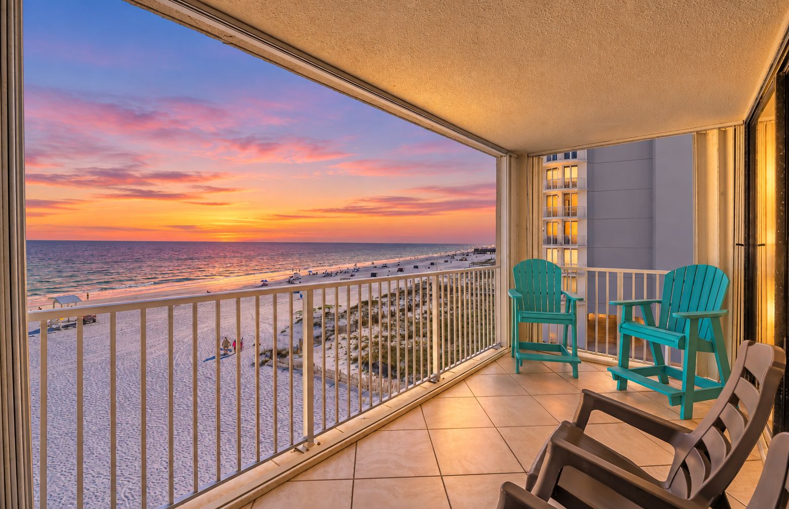 A balcony with a view of the beach at sunset, two chairs, and a small table.