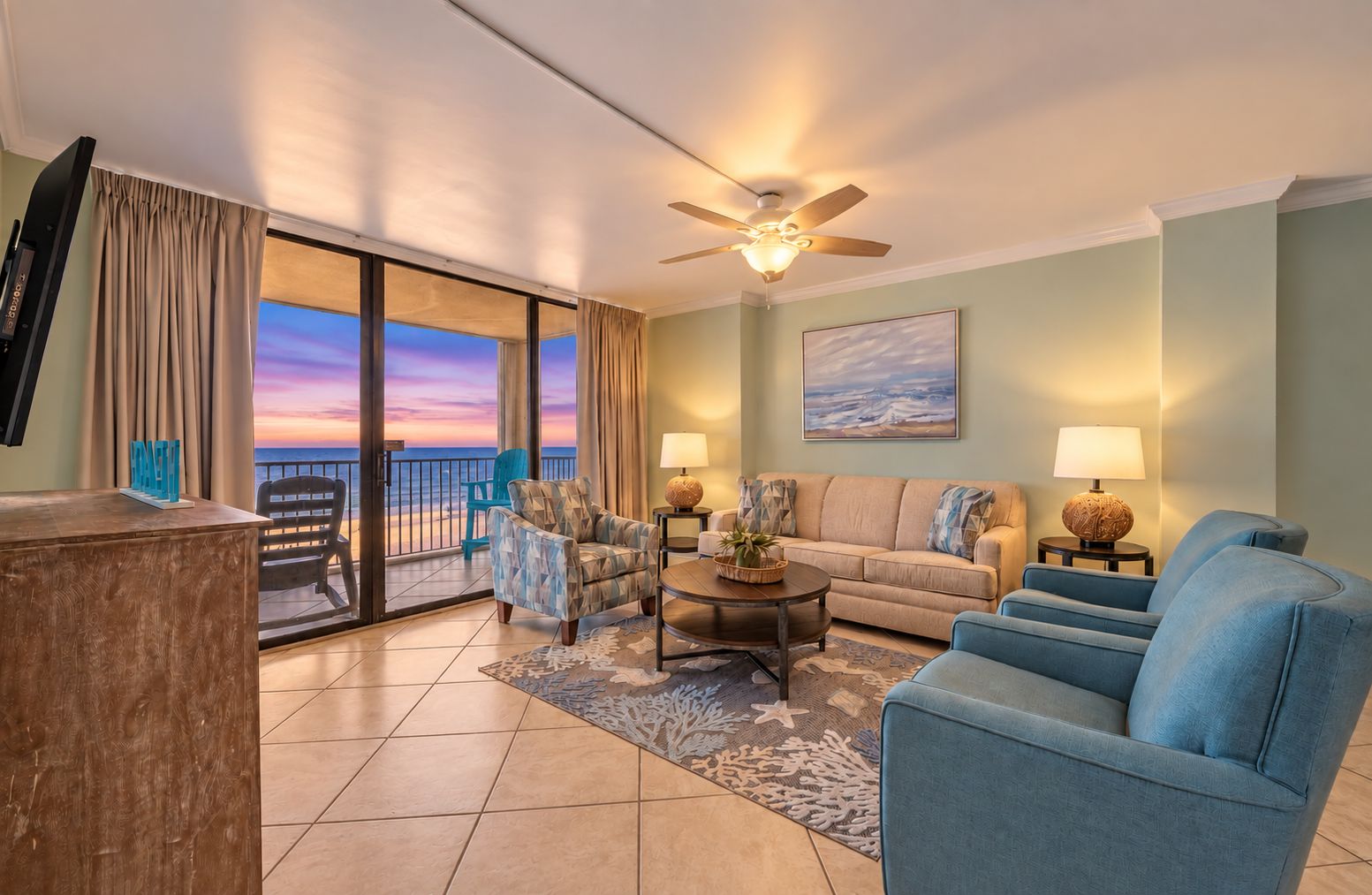 A living room with a sofa, armchairs, a coffee table, and a TV. There is a balcony with a view of the sunset.