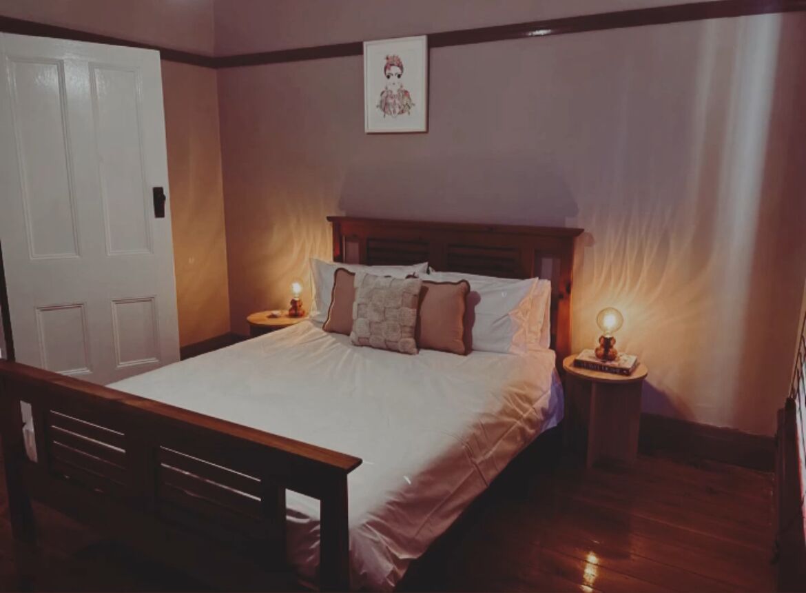 Een slaapkamer met een houten bed, wit beddengoed, twee nachtkastjes met lampen en een deur.