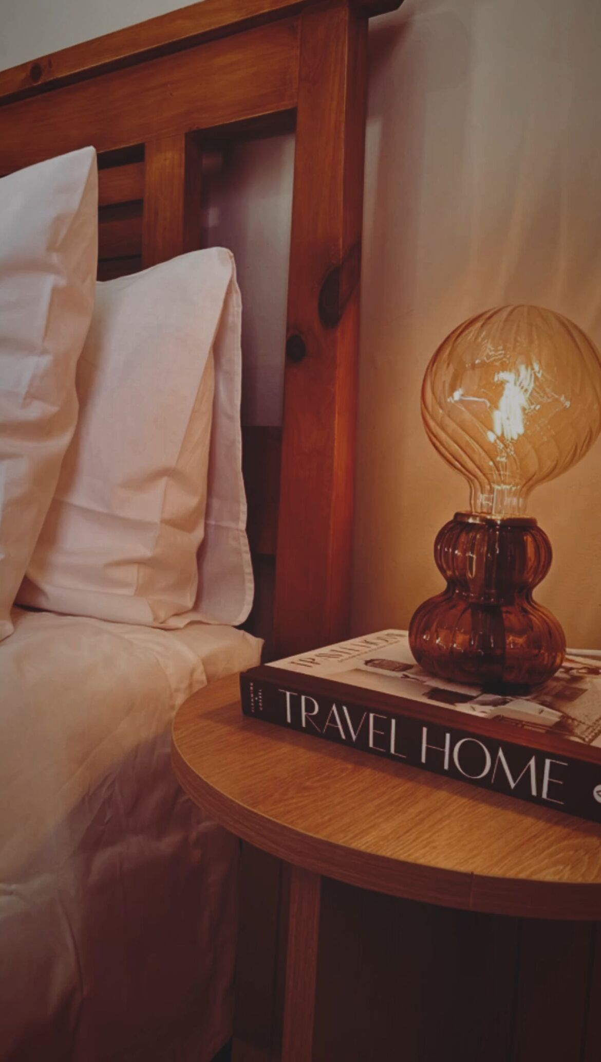 A neatly made bed with white linens, a round wooden side table with a book titled "TRAVEL HOME" and a decorative lamp.
