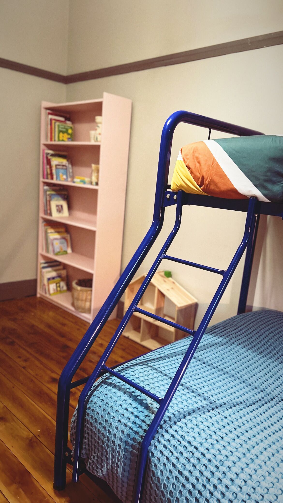 A bunk bed with a blue mattress and metal frame, a pink bookshelf filled with books, and a wooden floor.