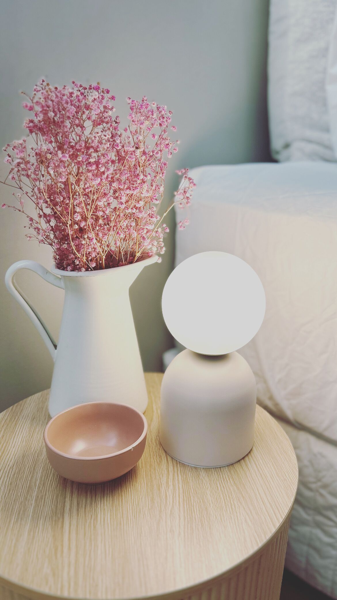 A white vase with pink flowers on a round wooden table, next to a white lamp and a pink bowl.
