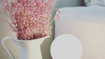 A white vase with pink flowers on a round wooden table, next to a white lamp and a pink bowl.