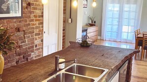 A kitchen with a brick wall, a wooden countertop, a stainless steel sink, hanging pendant lights, and a view to a dining area. - The BnB filled to the brim with character and charm! (Dubbo)