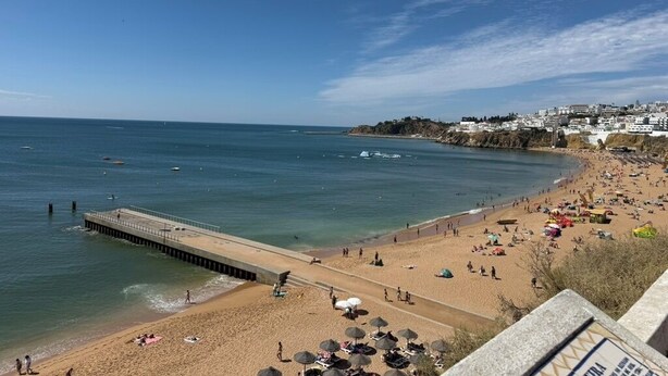 A beach with a pier extending into the water, people on the sand, and buildings in the background.