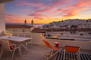 Una terraza con mesa y sillas, con vista a un pueblo costero al atardecer.
