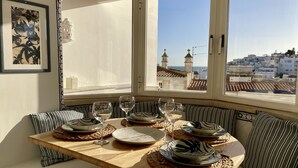A dining area with a wooden table set for four, surrounded by striped cushions, and a view of a coastal town through large windows.