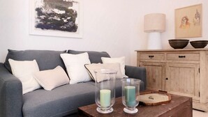 A living room with a grey sofa, white cushions, a wooden cabinet with two bowls, a lamp, and two glass candle holders on a wooden table. - Appartamento Cala Rotonda (Favignana)