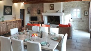Un comedor con una mesa grande dispuesta para seis personas, rodeada de sillas. Hay una chimenea con repisa sobre la mesa, y una pared de piedra al fondo.