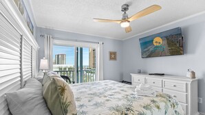 Una camera da letto con un letto, una ventola a soffitto, una TV a parete, una finestra con persiane e vista sugli edifici esterni.