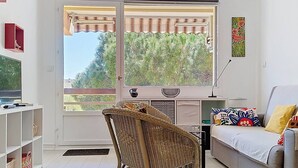 Een woonkamer met een bank, een rieten stoel, een televisie, een raam met uitzicht op bomen en een schilderij aan de muur.