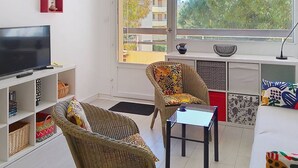 Een woonkamer met een televisie, rieten meubels, een glazen salontafel en uitzicht op gebouwen en bomen.