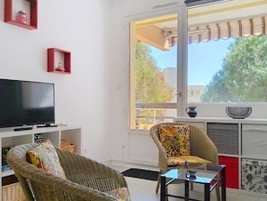 A living room with a television, wicker furniture, a glass coffee table, and a large window with a view of trees and buildings. - Airy 1BR Apt - AC, Loggia, Parking - Heart of La Favière (Bormes-les-Mimosas)