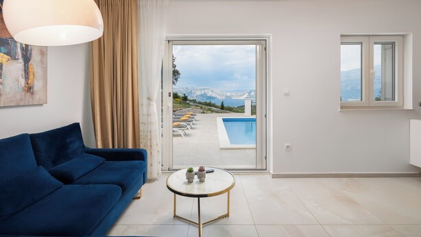 A modern living room with a blue sofa, a round coffee table, and a view of a pool and mountains through a glass door. - Emerald Eco Villa (Povlja)