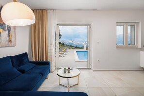Een moderne woonkamer met een blauwe bank, een ronde salontafel en uitzicht op een zwembad en bergen door een glazen deur.