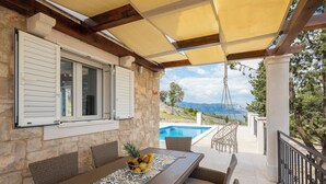 A covered outdoor dining area with stone walls, wicker furniture, and a view of a swimming pool and mountains. - Emerald Eco Villa (Povlja)