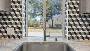 Een keuken met een rvs-gootsteen, een achterwand met een geometrisch patroon, en uitzicht op een gebied met bomen.