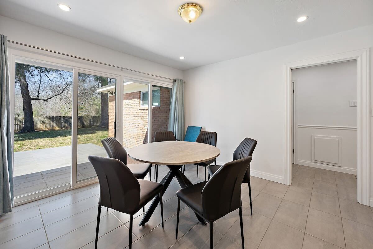 Un coin repas avec une table ronde et six chaises, de grandes portes-fenêtres coulissantes donnant sur un jardin, et une porte blanche sur le côté droit.
