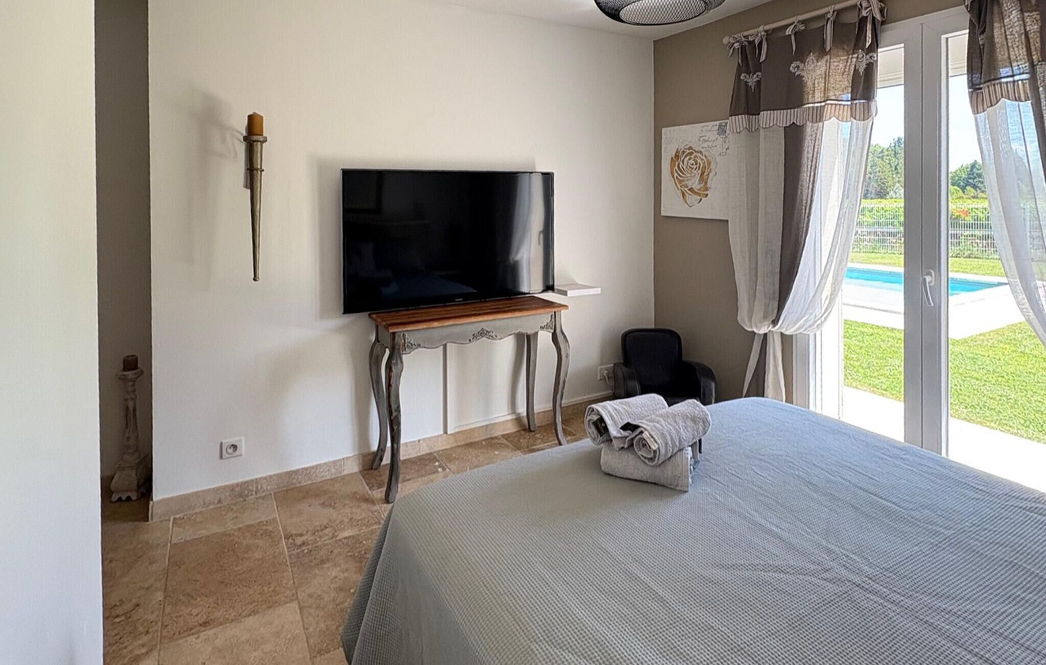 A bedroom with a bed, a television on a wooden stand, a chair, and a window with curtains.