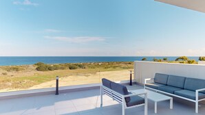 A balcony with a white sofa, a small table, and a chair, overlooking a beach and the sea. - Villa Ektoras at Periyiali Resort with  modern design, and amenities.  (Paralimni)