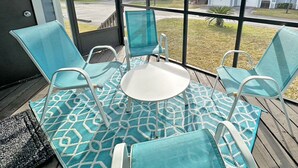 Un espace extérieur aménagé, abrité et doté d’une table et de quatre chaises, d’un tapis à motifs et d’une vue sur les maisons, visible à travers de grandes fenêtres.