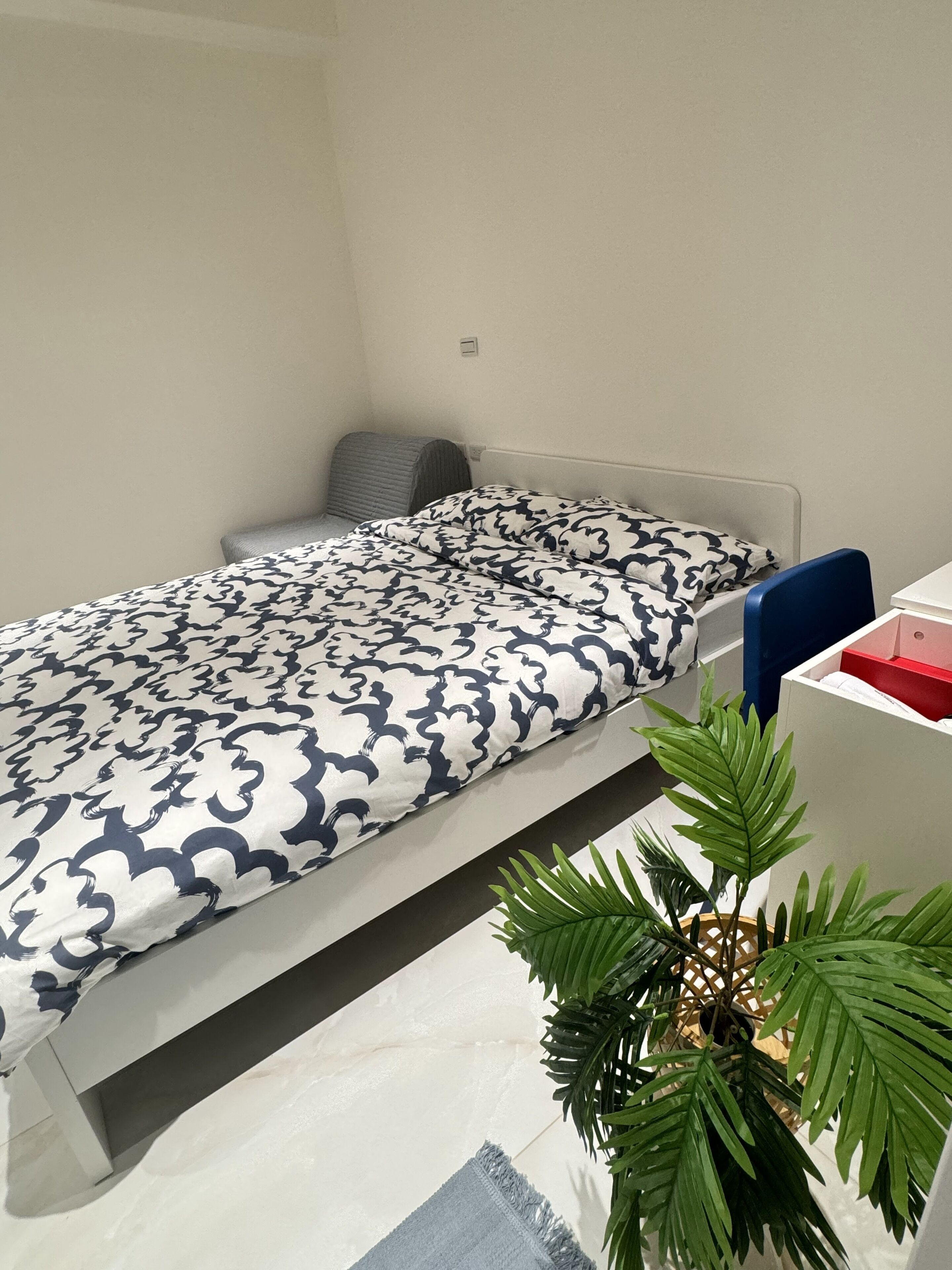 Una cama individual con un edredón estampado, una mesita de noche, una silla y una planta en maceta.