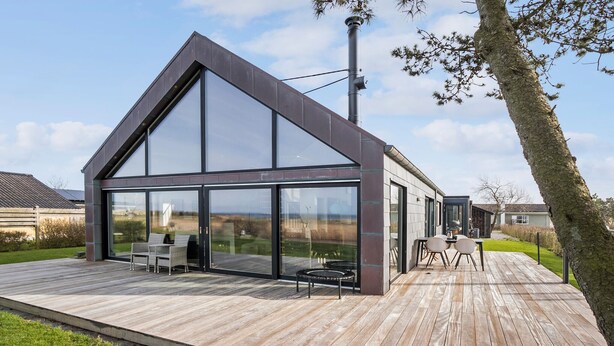 Una casa moderna amb una gran façana de vidre, una terrassa de fusta i mobiliari exterior.