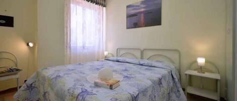Un dormitorio con cama, mesita de noche, cortinas en la ventana y un cuadro en la pared.