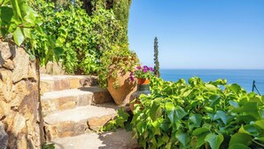 Eine steinerne Treppe führt zu einer Klippe mit Blick auf das Meer, umgeben von üppiger Vegetation und Topfpflanzen.