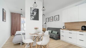 Un espacio de sala moderno y bien iluminado, con una mesa de comedor, sillas blancas, un sofá y una zona de cocina con armarios y encimera.