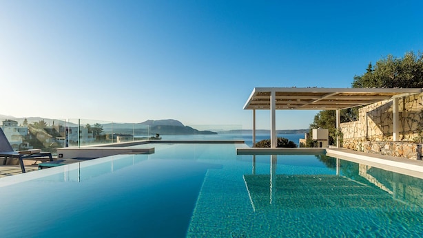 Uma piscina infinita azul cristalina com uma pérgola de um lado e uma parede de pedra do outro, com vista para uma paisagem costeira.