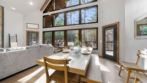 Un comedor con mesa de madera, sillas y sofá. Amplias ventanas ofrecen vistas a árboles.