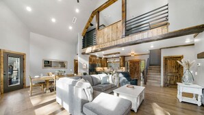 Un salón espacioso con un amplio sofá seccional gris, una mesa de centro blanca y una mesa de comedor con sillas. Una escalera de madera conduce a un nivel superior.