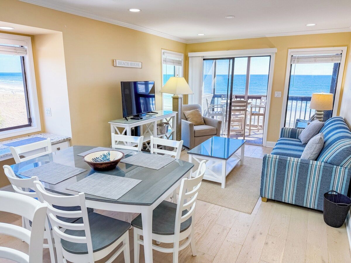A room with a dining table, chairs, a sofa, a TV, and a view of the beach.
