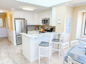 A modern kitchen with white cabinetry, a stainless steel refrigerator, a microwave, and a granite countertop island with bar stools.